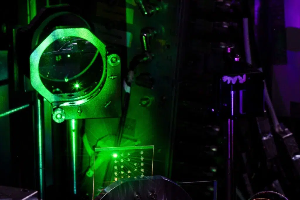 Dans une chambre à vide, une impulsion laser d’intensité relativiste est focalisée sur une cible en verre, générant une interaction extrême entre lumière et matière.Cette interaction produit un plasma lumineux verdâtre ainsi qu’un faisceau harmonique violet, caractéristique de la Coherent Harmonic Focus, capable de concentrer des champs lumineux cohérents d’une intensité exceptionnelle. Ces conditions ouvrent la voie à l’étude du vide quantique, où la lumière peut révéler des propriétés fondamentales de l’espace lui-même. (Crédit : Timmis et al., 2026)