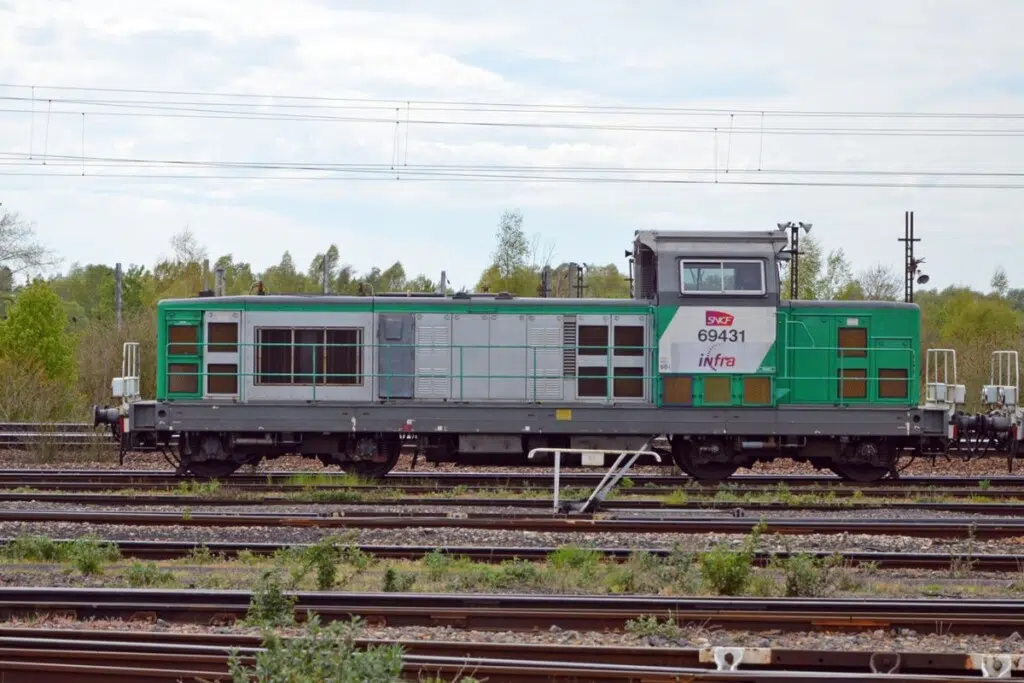 Les BB 69400 sont des locomotives diesel-électriques issues de la rénovation et de la remotorisation de 75 BB 66400 entre 2004 et 2009. Équipées d’un moteur MTU de 1 040 kW, elles développent 745 kW en continu et peuvent atteindre 120 km/h. Modernisées également au niveau de l’ergonomie, de l’insonorisation et de la climatisation de la cabine, elles sont notamment utilisées par SNCF Infra ; 60 exemplaires étaient encore recensés fin août 2026 (crédit : BB 69431 : depot de longueau.jpg de Wikimedia Commons par BB 22385 , CC-BY-SA 4.0)